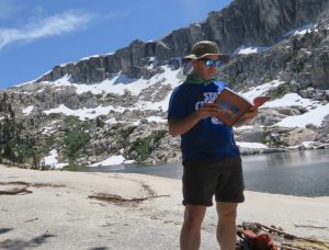A man standing and reading a book next to a lake and a snow-spotted mountain