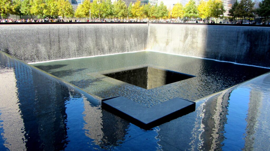 Water flows down two walls and disappears into a void at the center of a memorial reflecting pool. Sunlight creates shadows on the pooled water.