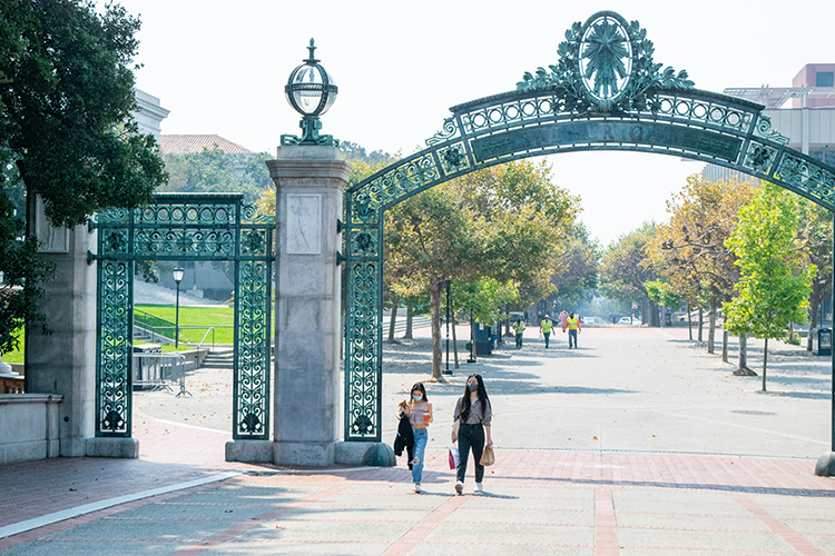 Two students in masks walk under Sather gate