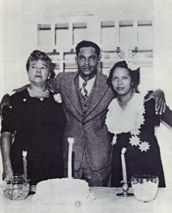 Two women and one man standing in front of a table with arms around each other, looking into camera.