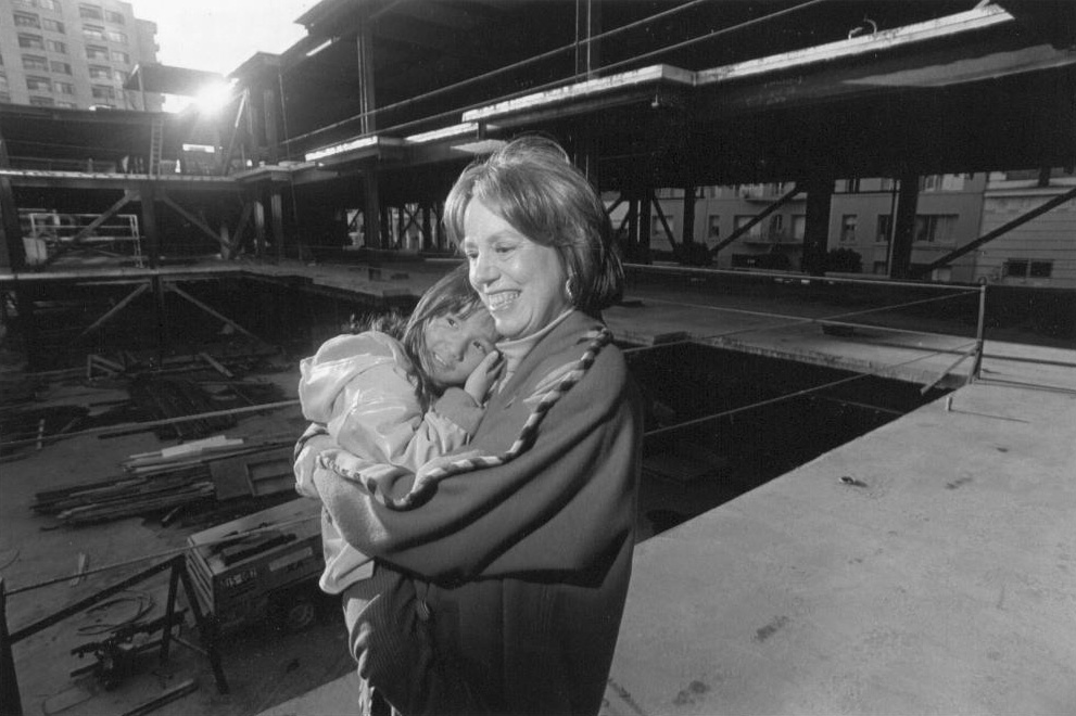 Midge Wilson holding a girl, against the backdrop of a school building during construction.