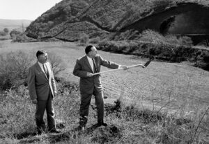 Harvey Banks stands near Governor Brown, who has a shovel. Both are wearing suits and standing amidst shrubs, with a valley and hill in the background. 