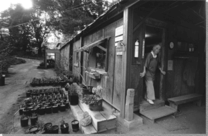 Gerda Isenberg coming out of a doorway of a small wooden building. There’s a sign that says “office,” and plants in pots along the side of the building.