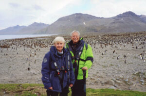 Two people in thick coats stand on a beach with thousands of penguins and mountains in the distance 