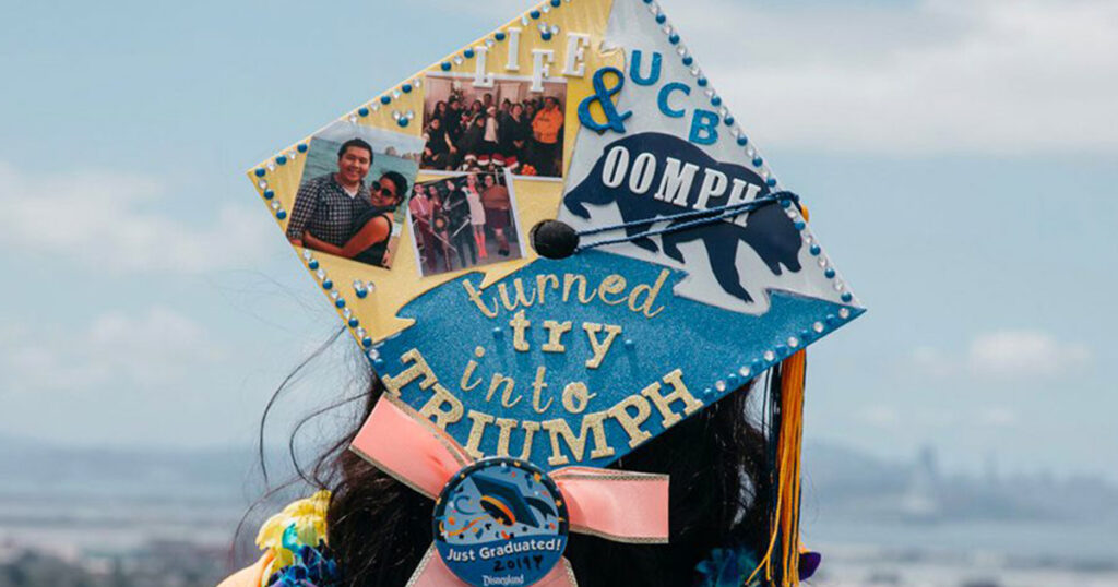 Close-up of the back of a decorated graduation cap on a person with long hair; the cap says, “Life, UCB, and OOMPH turned try into triumph.” At the bottom of the cap, there is a button fastened to a bow; the button depicts a graduation cap surrounded by confetti, and says “just graduated!” 