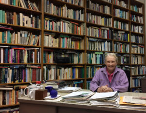 Color photograph of Carolyn Merchant seated in her office on campus behind a desk covered with stacks of paper, which sits in front of a floor-to-ceiling bookshelf entirely full of books