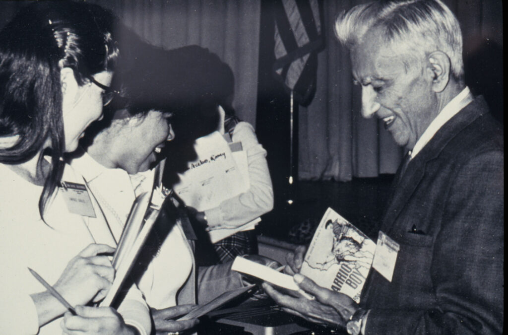 An older Ernesto Galarza at a book signing, signing a book, with two young women waiting in line; everyone is smiling.
