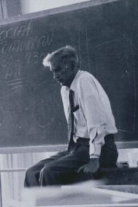 Ernesto Galarza sitting on a desk, with chalk board in the background, looking serious.