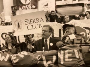 Black and white photograph of Robert Cox wearing a coat and tie and speaking into a microphone while surrounded by environmentalists who hold Sierra Club signs 