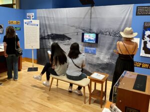 Several people sit and stand in the exhibit space, with some sitting in front of a video display while others stand and look at photographs on the wall of the exhibit