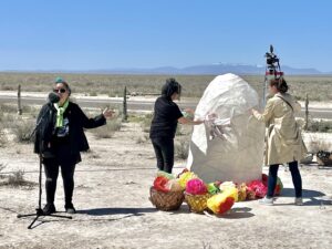In the desert a woman stands at a microphone and two women stand next to an art installation of a large rock
