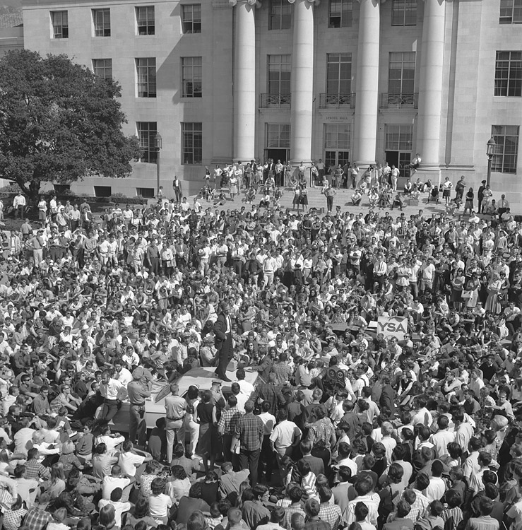 A young woman stands on top of a police car in the middle of Sproul Plaza. She is surrounded by a large crowd. 